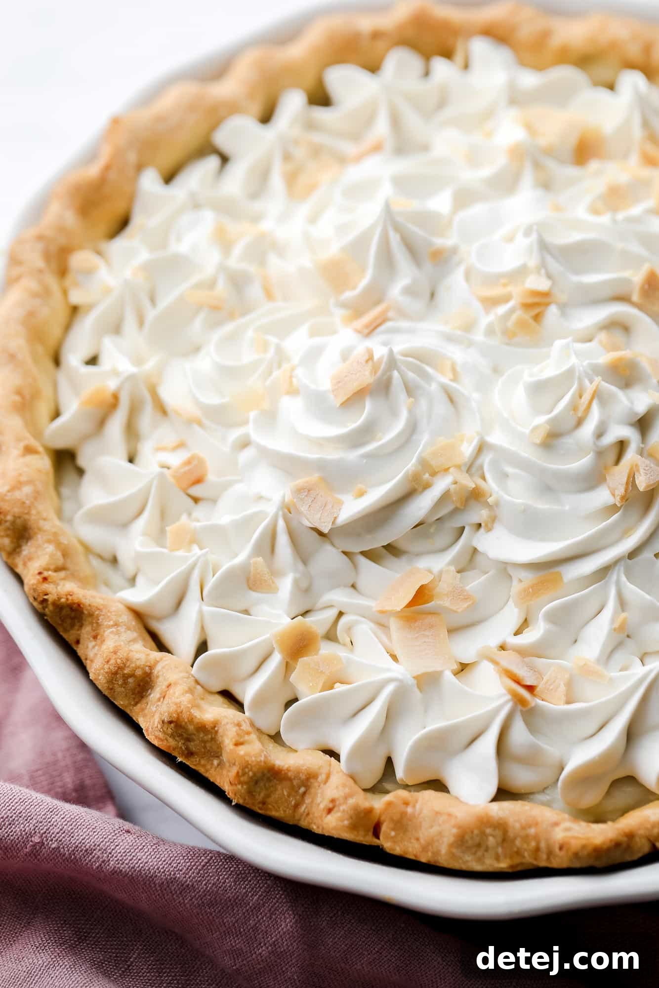 A close-up view of a beautifully textured vegan coconut cream pie, topped with swirls of whipped cream and delicate coconut shreds.