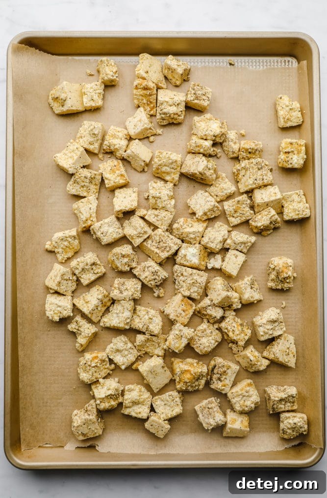 Close-up of ripped tofu chunks seasoned with a light coating, spread evenly on a parchment-lined baking pan, ready for baking to achieve crispiness.