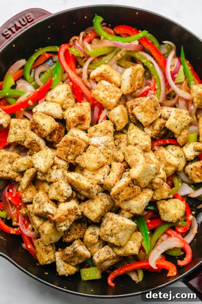 Golden, cooked ripped tofu pieces are added to a large skillet with sautéed bell peppers and onions, ready to be combined with the sauce.