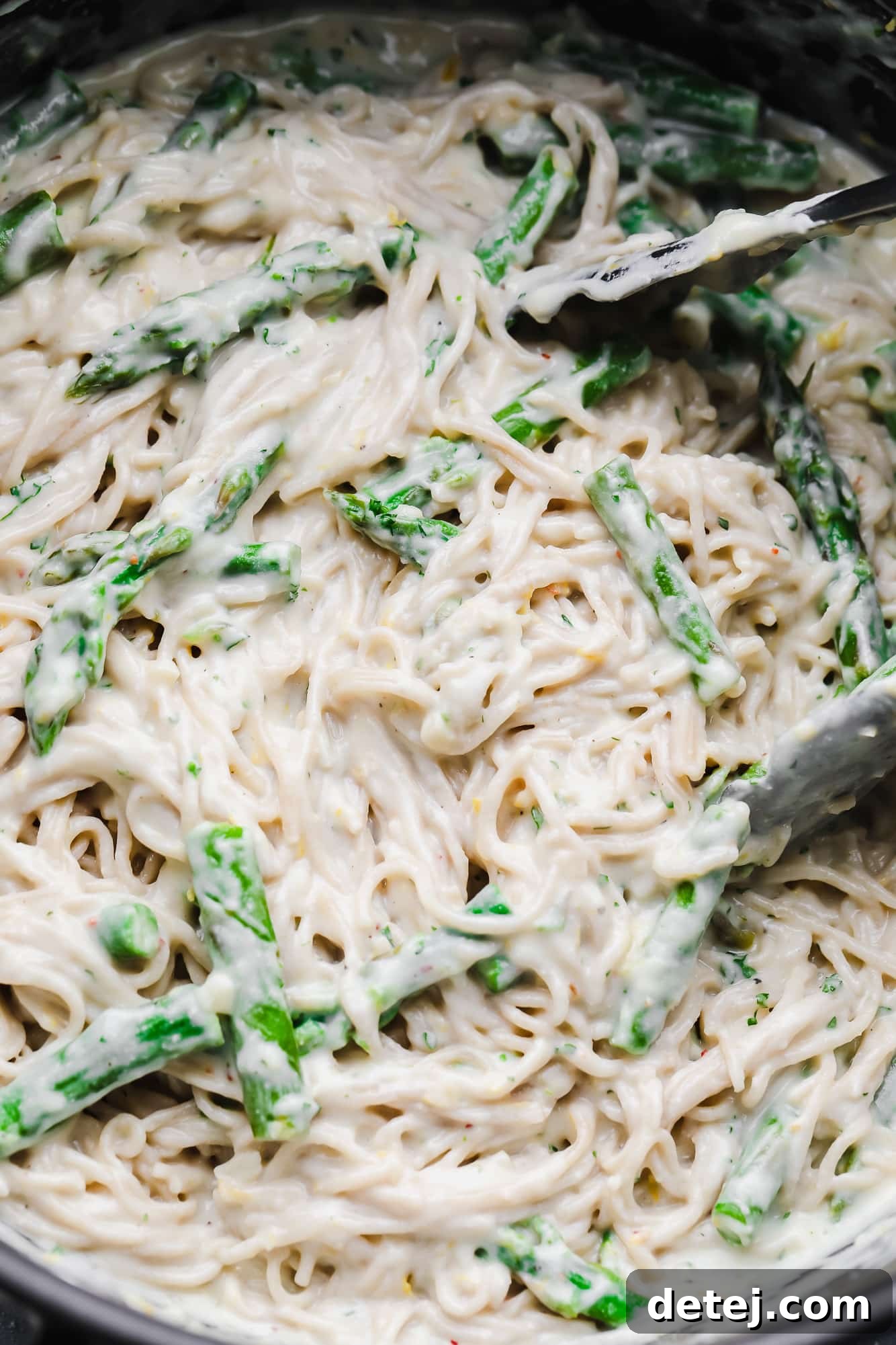 Tossing lemon asparagus pasta with tongs in a large black pot, coated in a creamy sauce.