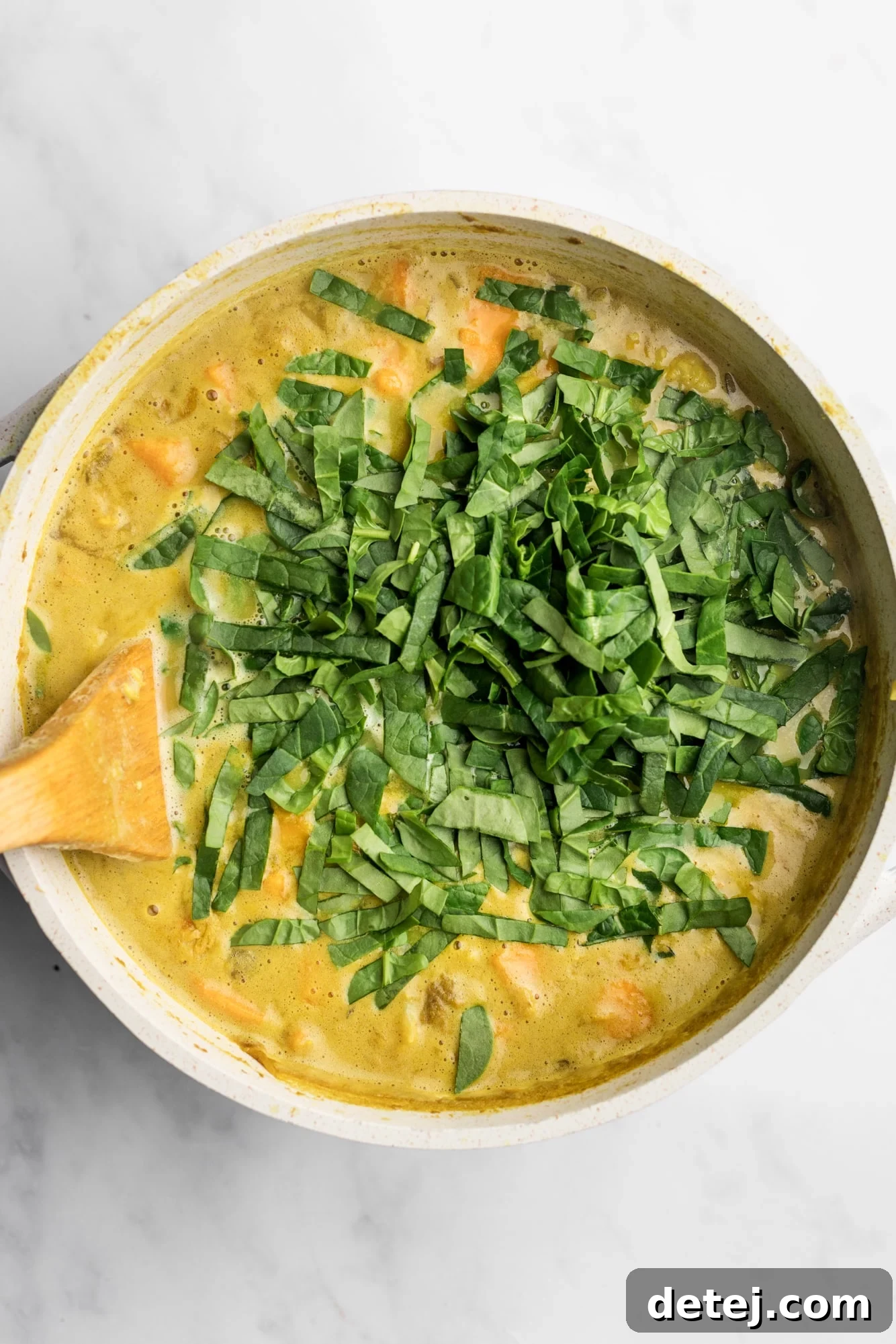 A wooden spoon stirring fresh baby spinach into a large pot of simmering coconut curry lentil soup.