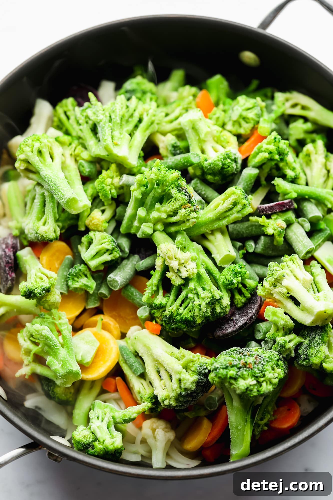 A generous portion of frozen mixed vegetables being cooked in a large black skillet, steam visibly rising as they thaw and sizzle.