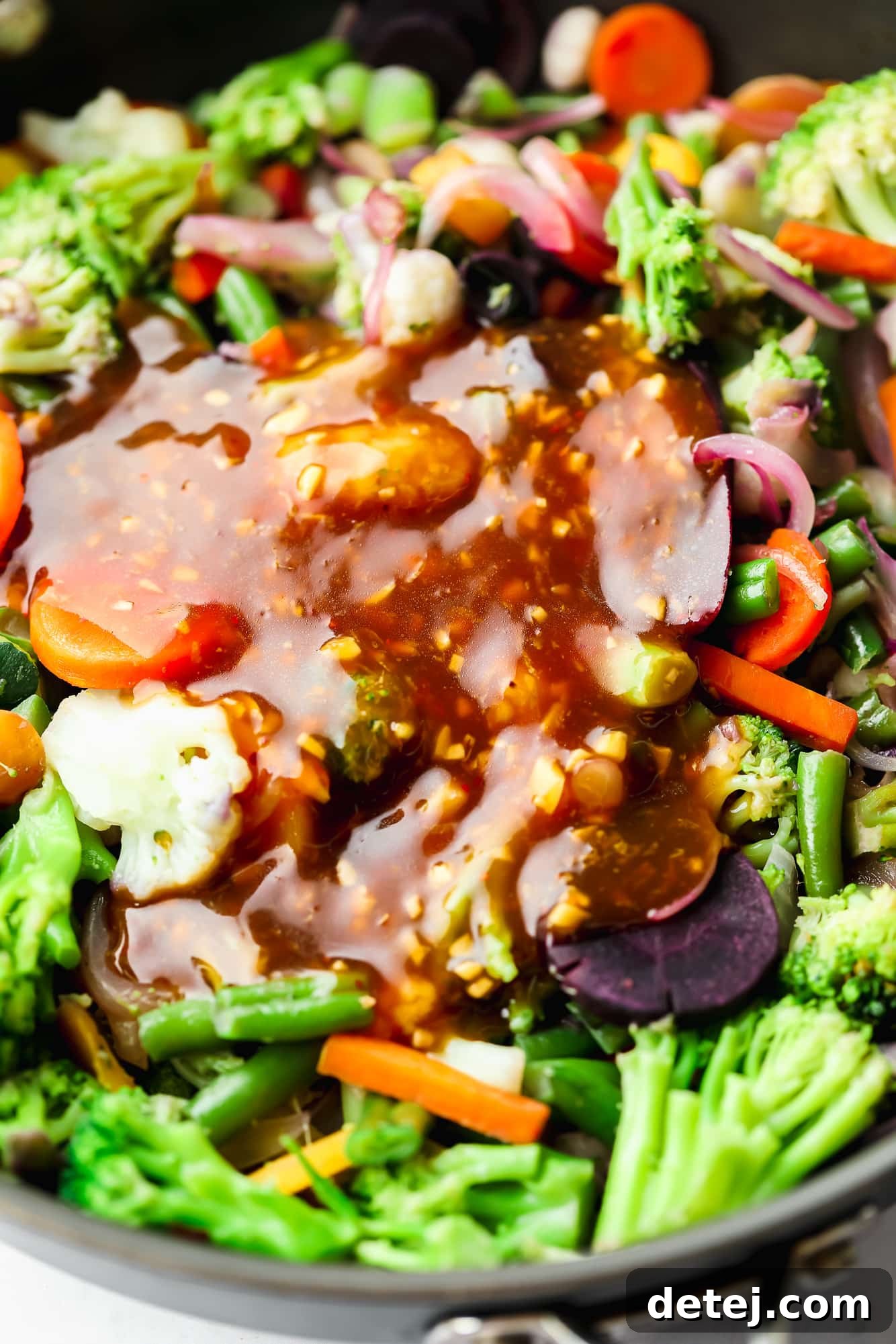 A rich, glossy stir fry sauce being poured over a colorful assortment of cooking vegetables in a black skillet, creating a delicious sheen.