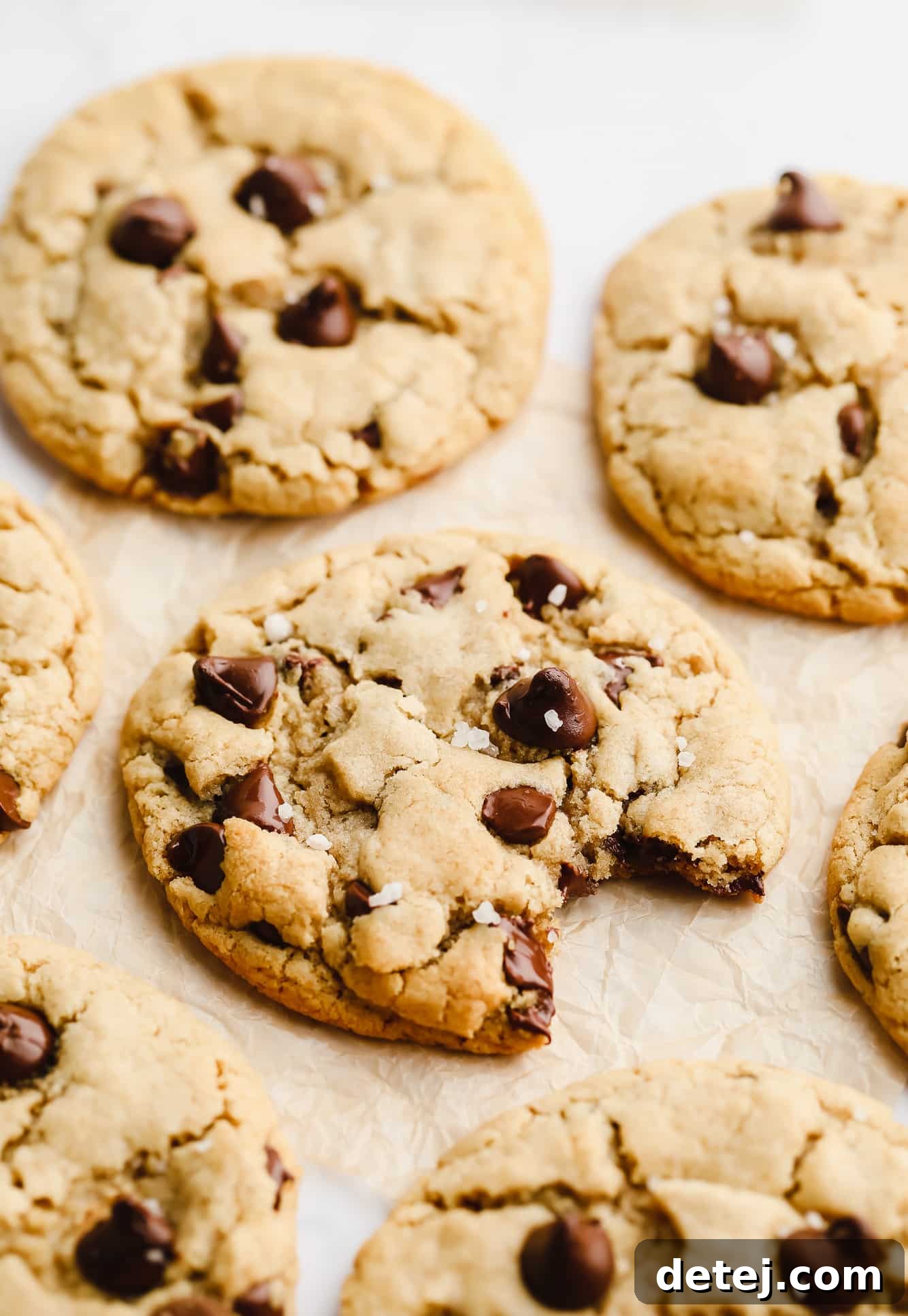 A freshly baked vegan gluten-free chocolate chip cookie, lightly sprinkled with sea salt, resting on parchment paper. Other cookies are blurred in the background, hinting at a delicious batch.