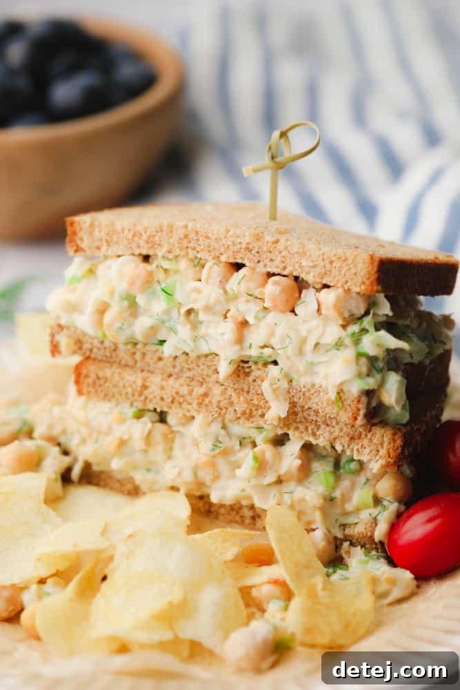 Close-up of a delicious chickpea salad sandwich on whole wheat bread, with a bowl of fresh blueberries in the blurred background. The sandwich is generously filled and garnished with fresh greens.