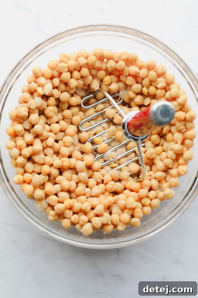 A close-up shot of chickpeas being mashed in a white bowl with a silver potato masher, showing a satisfying chunky texture.