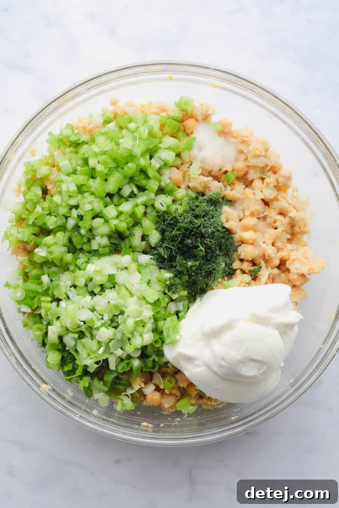 A mixing bowl filled with mashed chickpeas, alongside vibrant green chopped celery, green onions, and fresh dill, with creamy vegan mayonnaise ready to be stirred in.