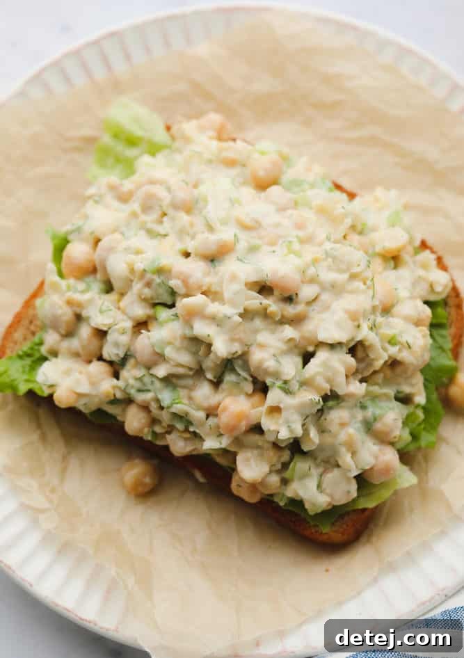 A close-up of a perfectly assembled chickpea salad sandwich on toasted bread, topped with fresh lettuce, showcasing the creamy chickpea mixture. The background is softly blurred.