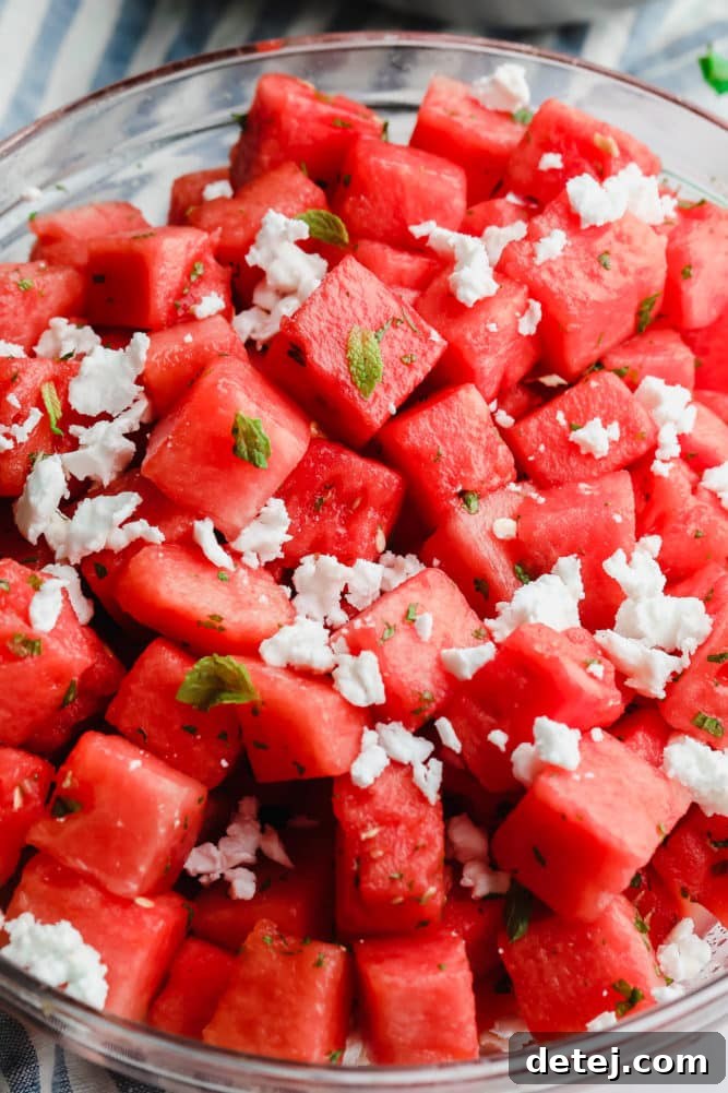 A beautifully presented glass bowl of watermelon feta salad, garnished with mint, beside a stylish blue striped towel on a marble surface.