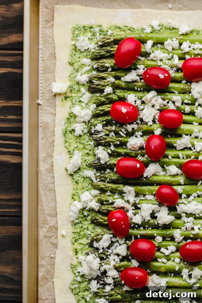 Unbaked asparagus puff pastry tart with pesto, fresh asparagus, cherry tomatoes, and crumbled vegan feta cheese on a baking pan, ready for the oven.