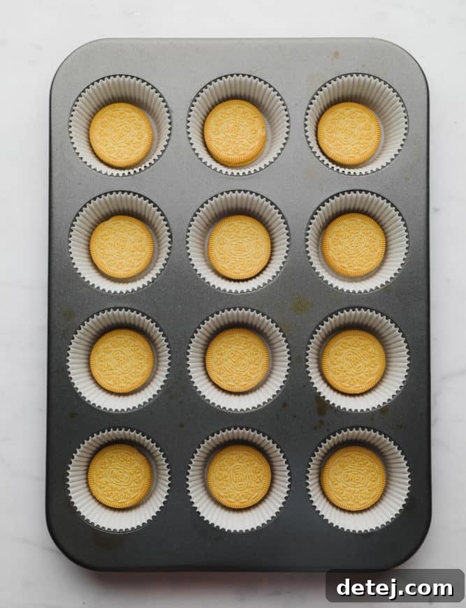 A 12-cup muffin pan neatly lined with paper wrappers, each holding a bright yellow lemon Oreo cookie at its base, ready for the filling.