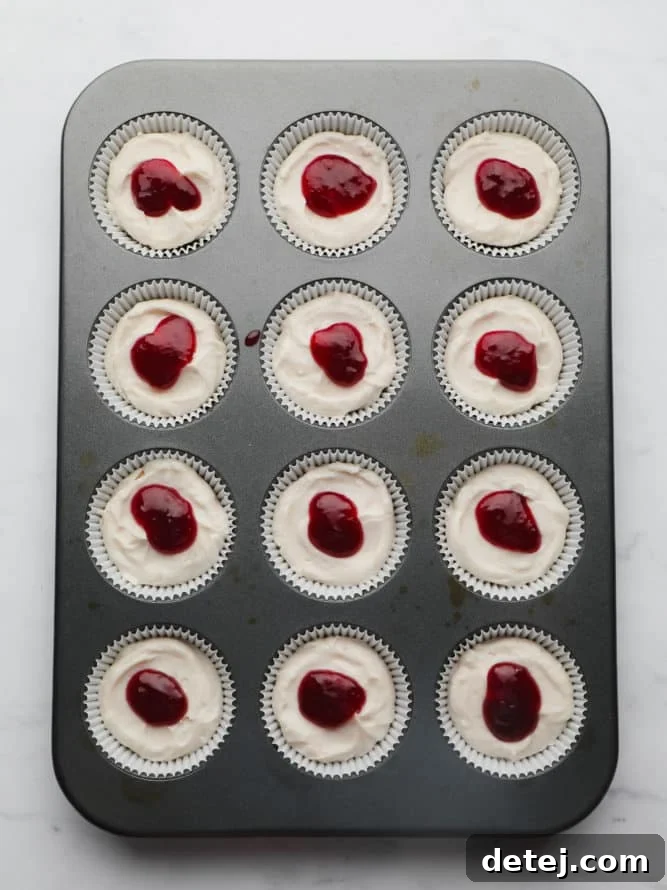 Close-up of a muffin pan showing individual cheesecake cups, each topped with a dollop of raspberry jam and a beautiful swirl pattern created with a toothpick.