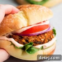 A square image of a hand reaching to grab a juicy vegan sweet potato black bean burger, served on a bun with fresh toppings.