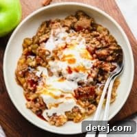 Scoops of warm vegan apple crumble, elegantly topped with vanilla ice cream, served in a white bowl with two spoons, ready to be shared.
