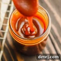A glass jar with rich, golden vegan salted caramel sauce gracefully drizzling into it from a spoon, illustrating its perfect consistency.