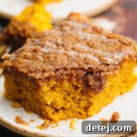 A close-up of a square slice of rich, orange vegan pumpkin coffee cake on a pristine white plate, showcasing its streusel topping.