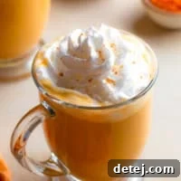 A square image of a warm vegan pumpkin spice latte in a glass cup, topped generously with whipped cream and a sprinkle of cinnamon.