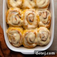 A square image displaying warm vegan pumpkin cinnamon rolls nestled in a baking dish, generously topped with maple cream cheese icing.
