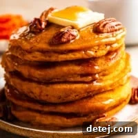 A close-up of a tall stack of vegan pumpkin pancakes, beautifully adorned with maple syrup, toasted pecans, and a dollop of vegan butter.