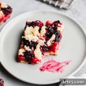 Square image of a delicious vegan blackberry bar with a shortbread crumble topping and a small bite taken out, presented on a grey plate.