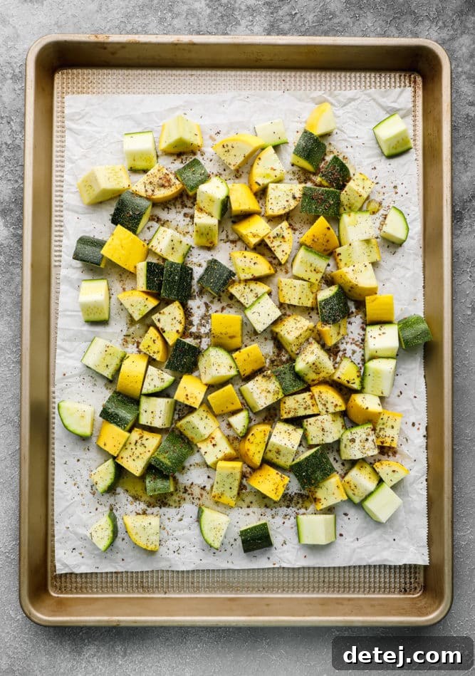 Parchment-lined gold pan with uncooked chunks of yellow squash and zucchini with seasonings on a grey background