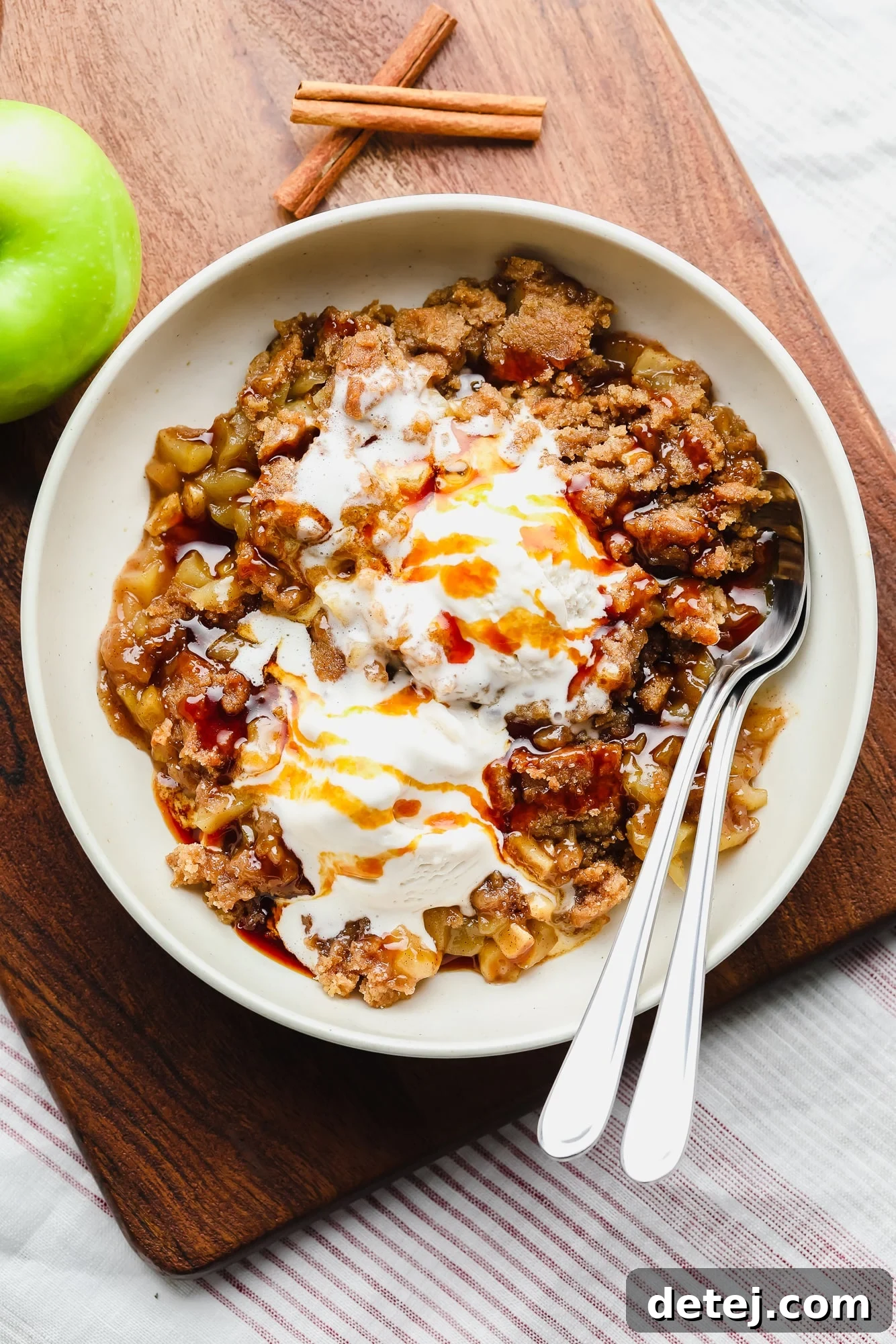 Scoops of golden-brown vegan apple crumble with melting vanilla ice cream in a white bowl, ready to be enjoyed with two spoons.
