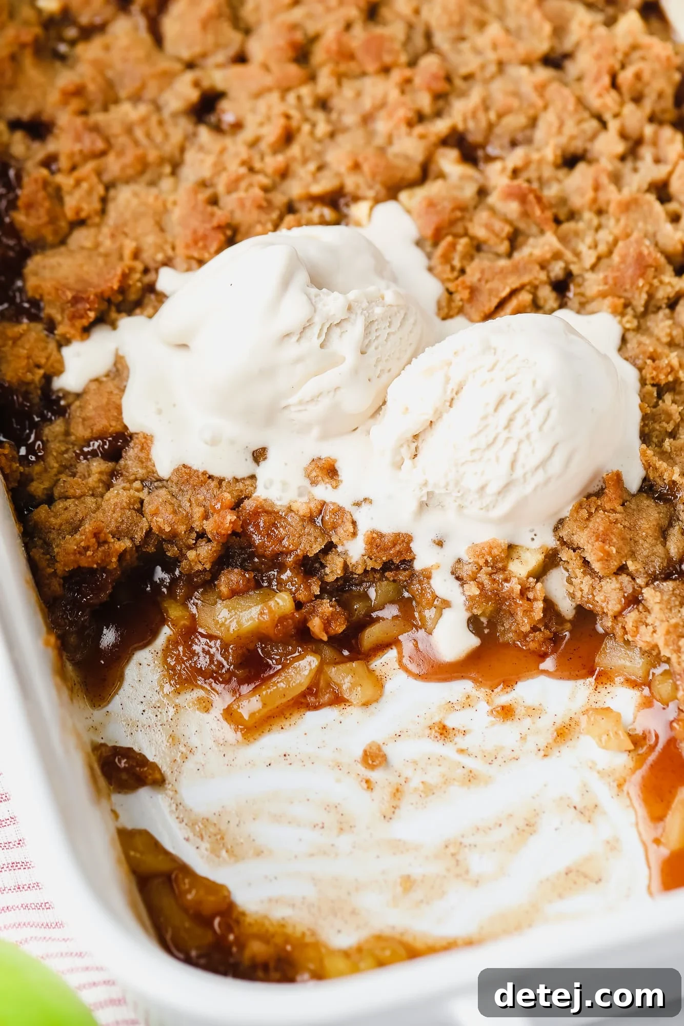 Close-up shot of baked vegan apple crumble in a white casserole dish, with one scoop missing, topped with melting vegan vanilla ice cream.