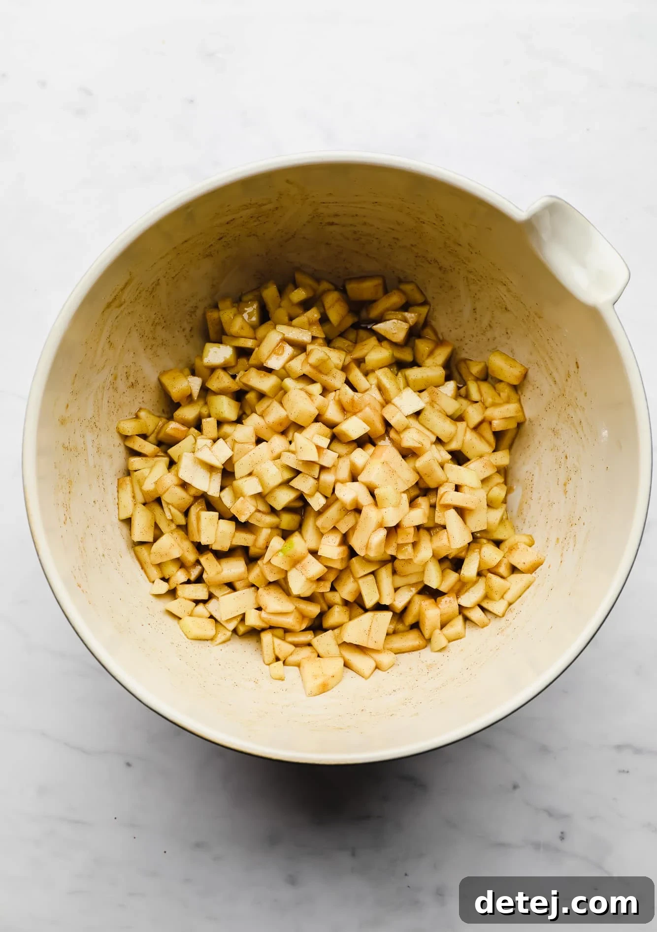 Diced apple chunks coated generously in cinnamon and brown sugar, ready for baking in a large white mixing bowl.