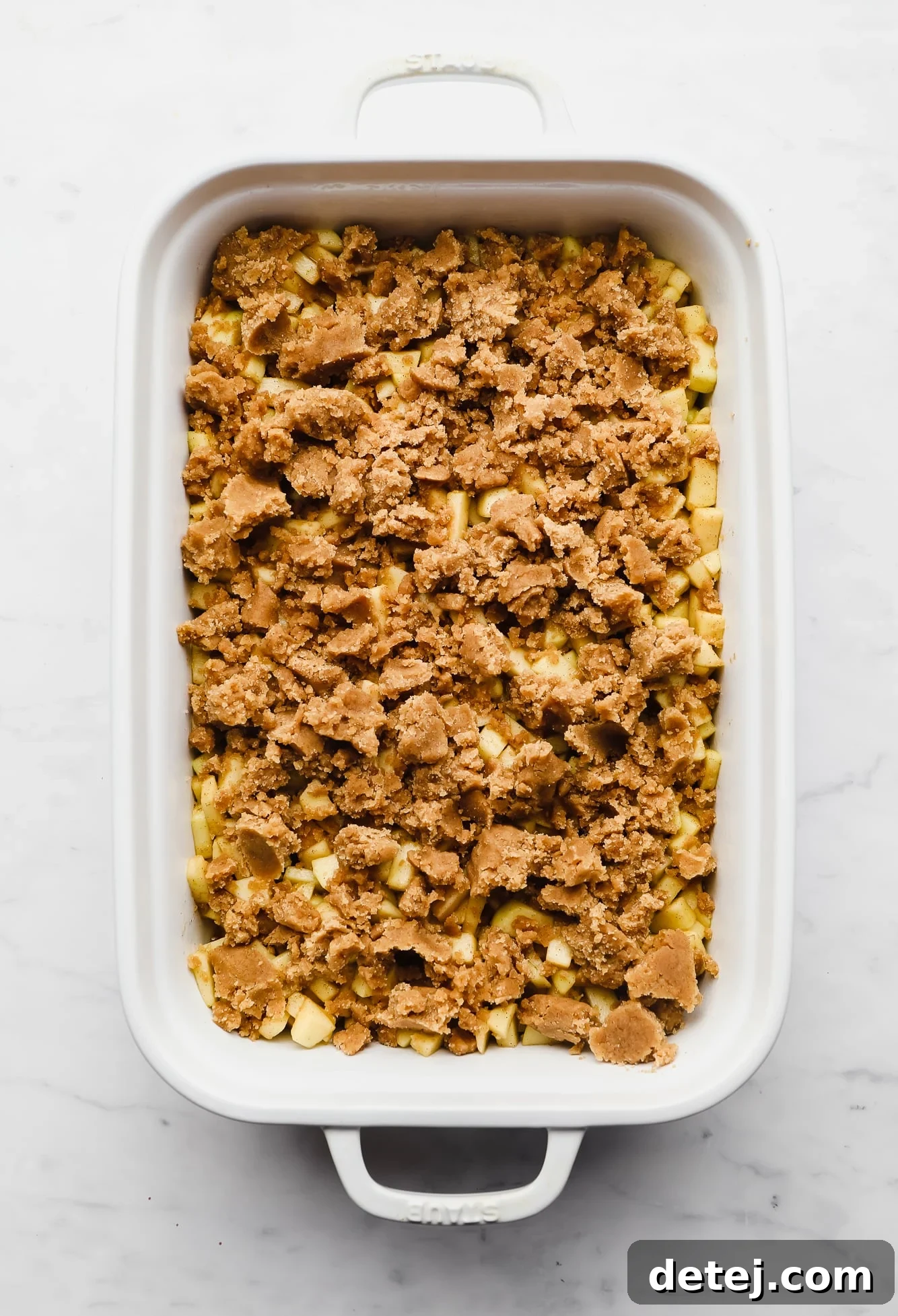 Unbaked vegan apple crumble in a large white casserole dish, with a generous layer of streusel topping over the cinnamon apples.