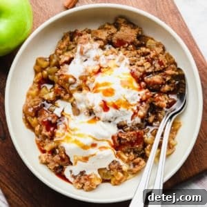 Scoops of vegan apple crumble with vanilla ice cream on top in a white bowl with 2 spoons.
