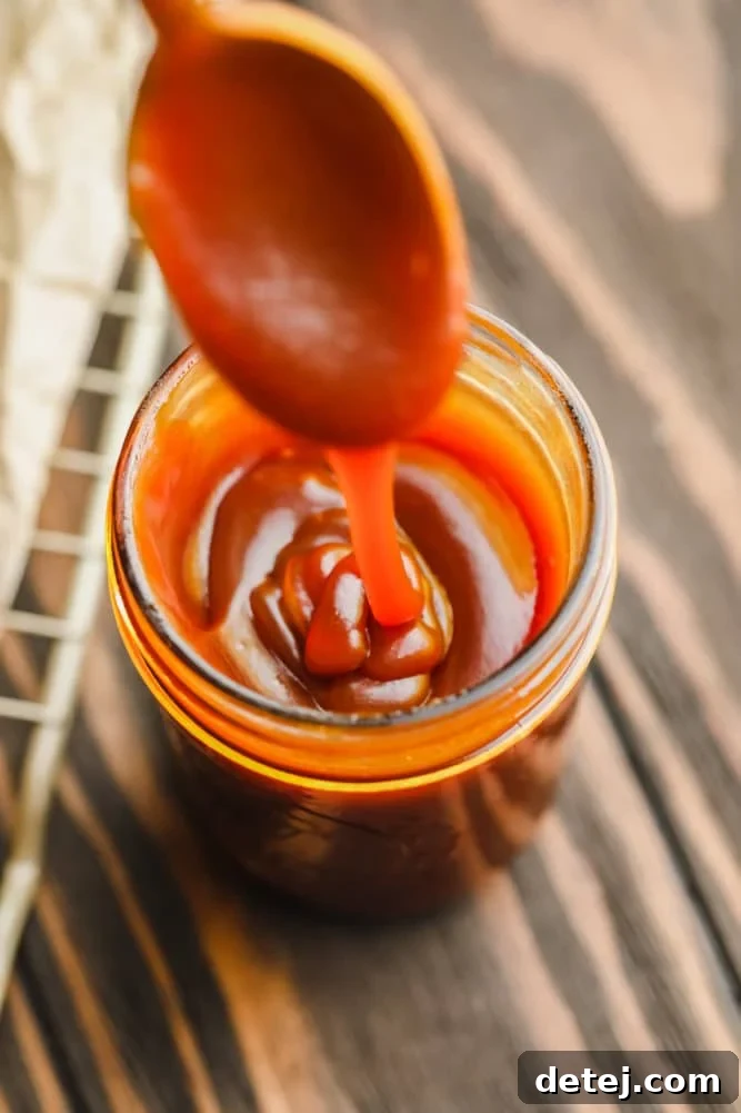 A glass jar filled with luscious caramel sauce, with a spoon drizzling more caramel into it. The backdrop is a rustic wooden surface, emphasizing homemade goodness.