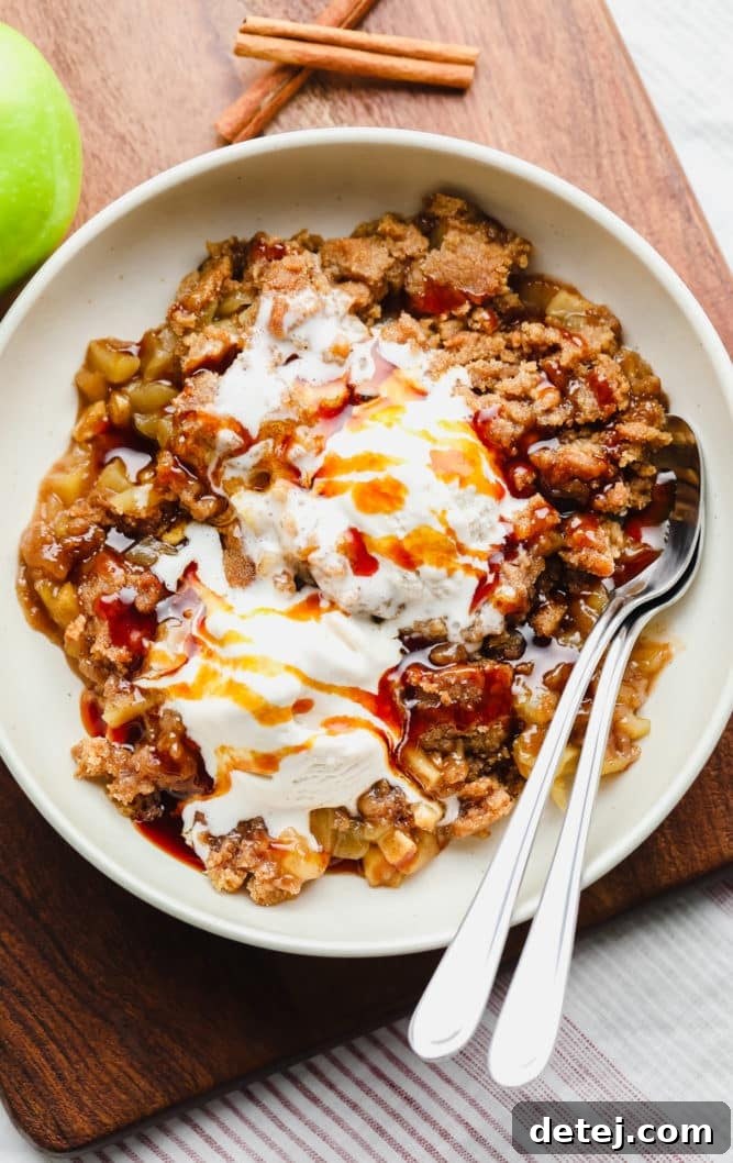 A rustic bowl of apple crisp topped generously with vegan salted caramel sauce and a scoop of non-dairy ice cream, with two spoons ready for indulgence on a wooden board.