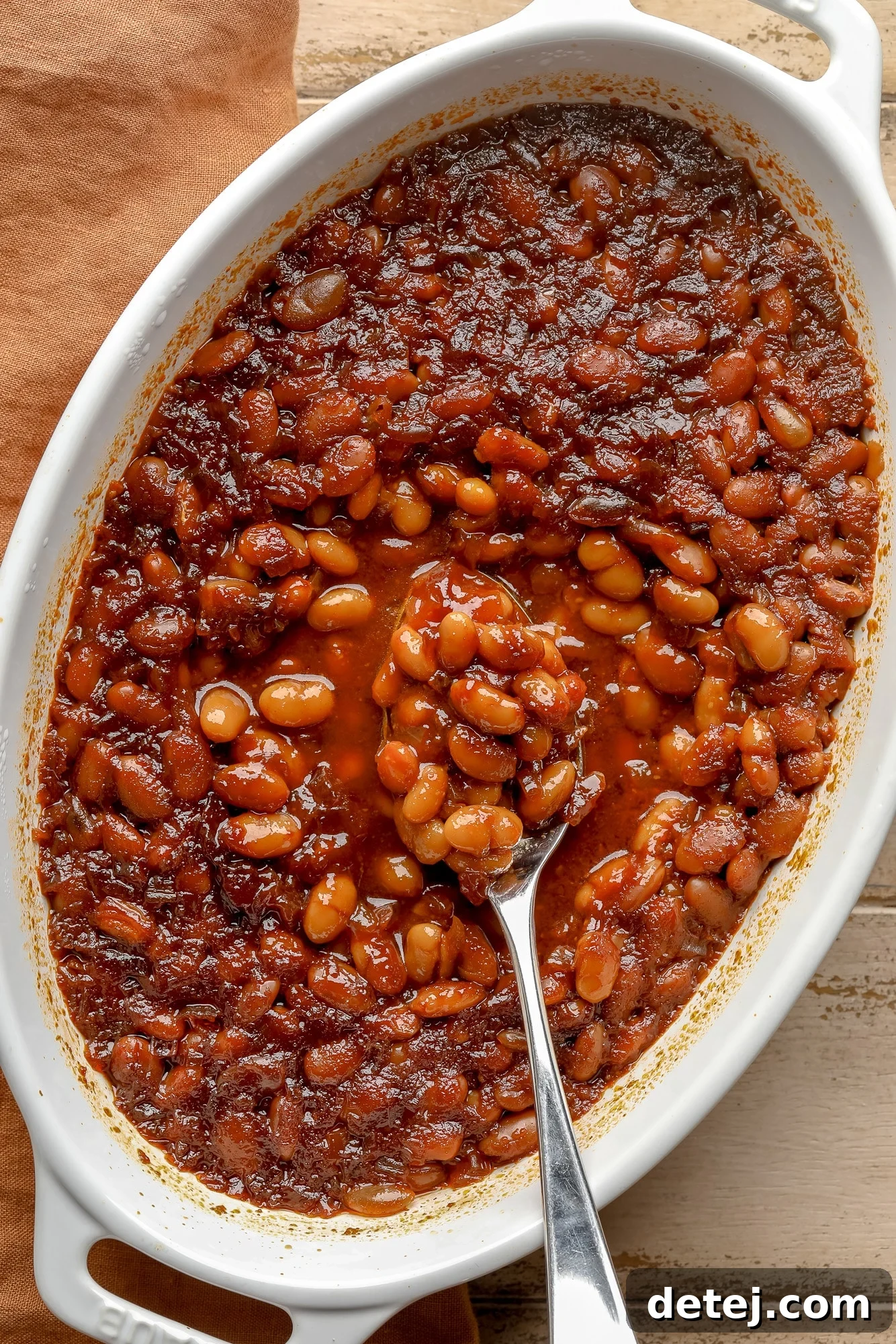 A serving spoon lifting a spoonful of rich, saucy vegan baked beans from a rustic casserole dish, showcasing the caramelized top and tender beans.