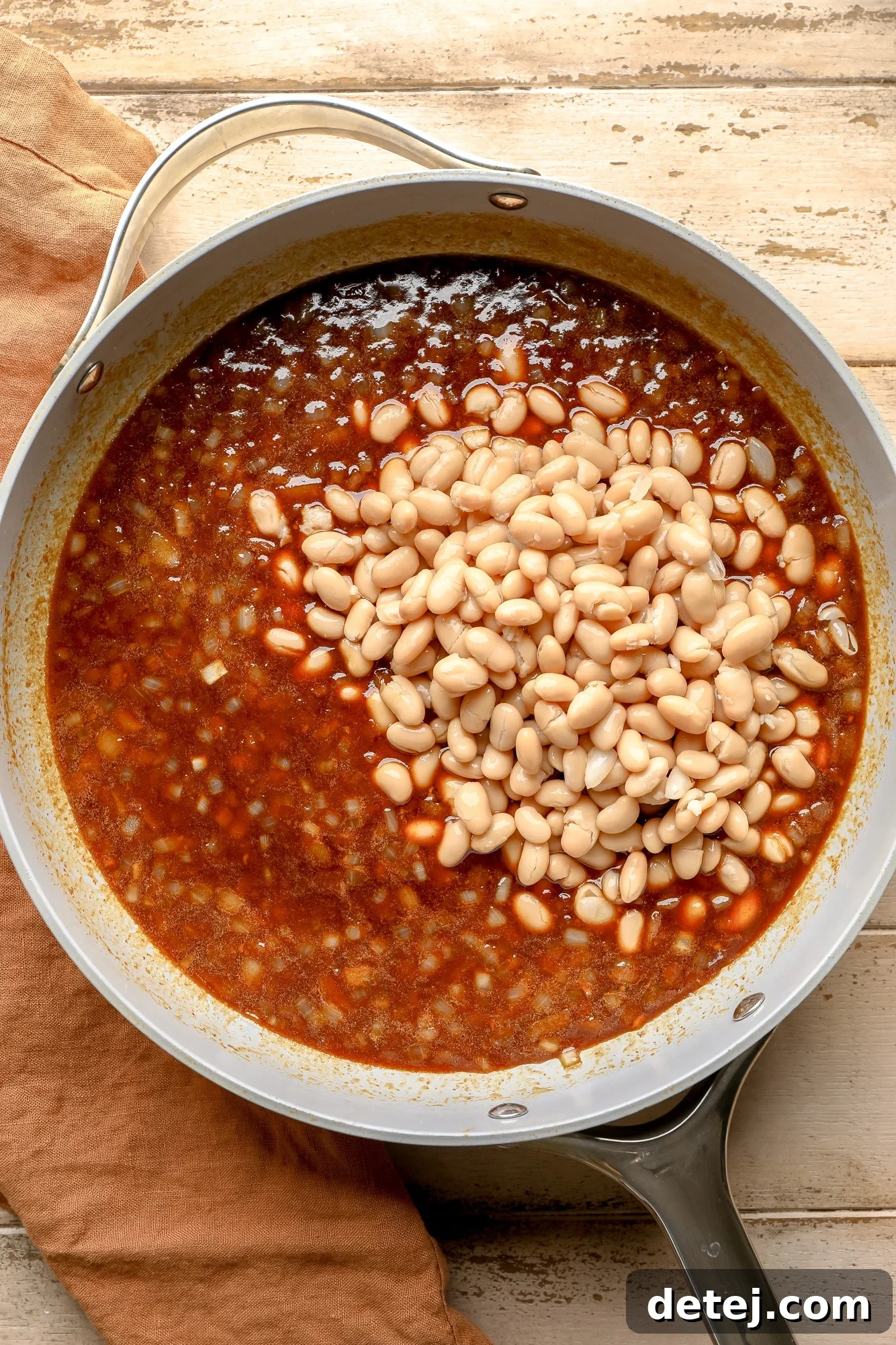 Creamy white navy beans are gently stirred into a rich, reddish-brown baked bean sauce in a large skillet, ensuring every bean is coated.