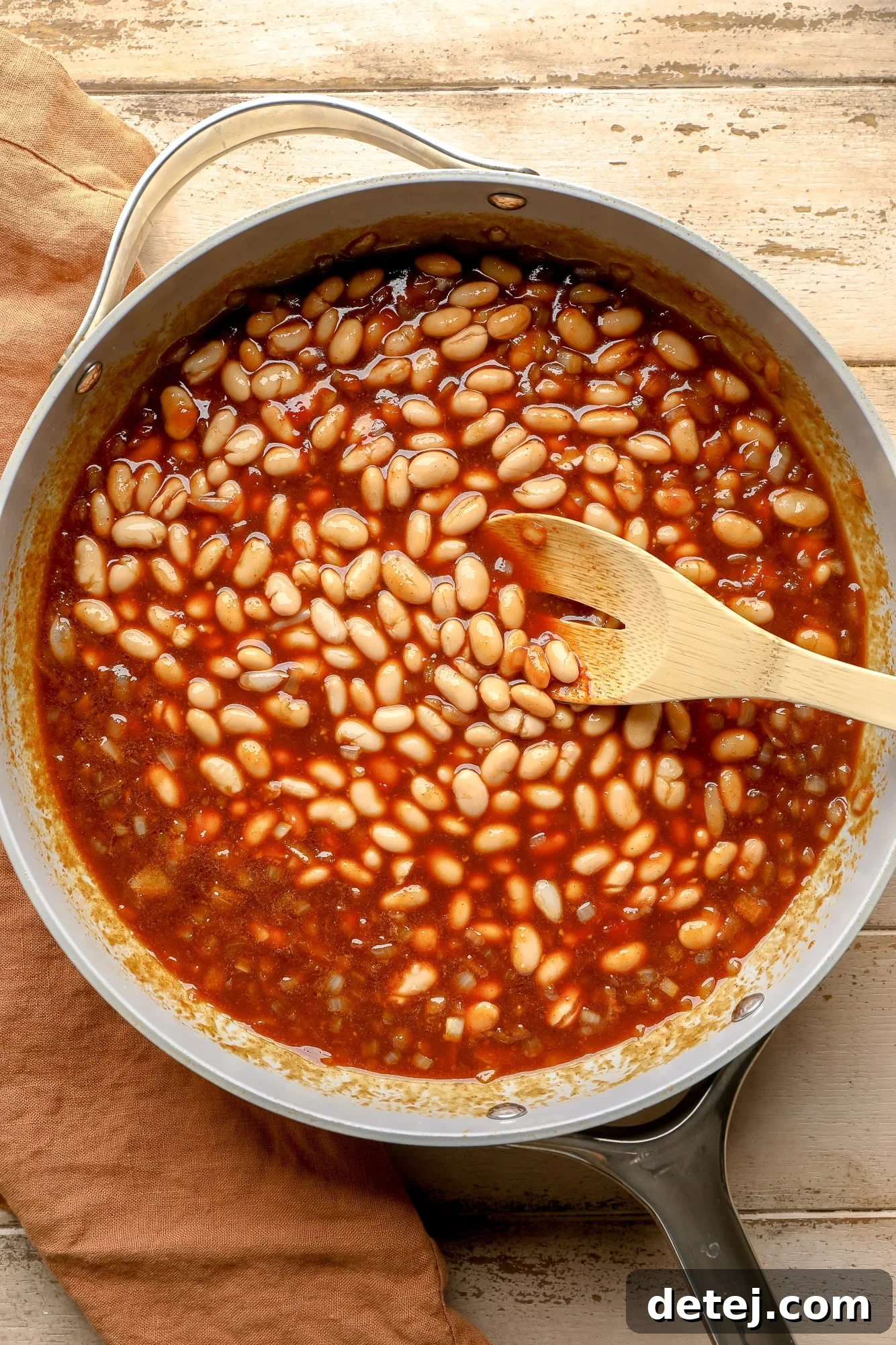 A wooden spoon expertly mixes tender white beans into a thick, sweet, and smoky sauce, ensuring even coating before baking.