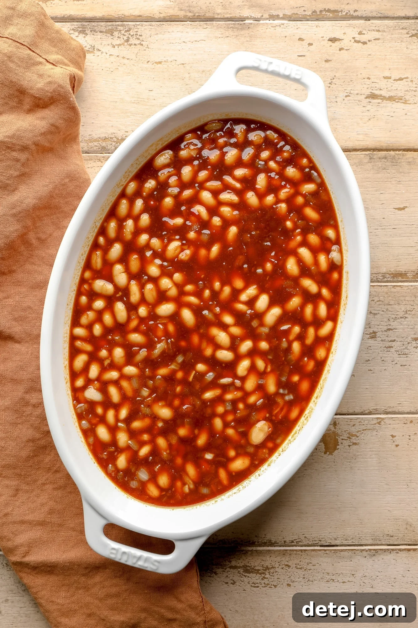 A large casserole dish filled with uncooked vegan baked beans, their creamy white beans glistening under a rich, thick red sauce, ready for the oven.