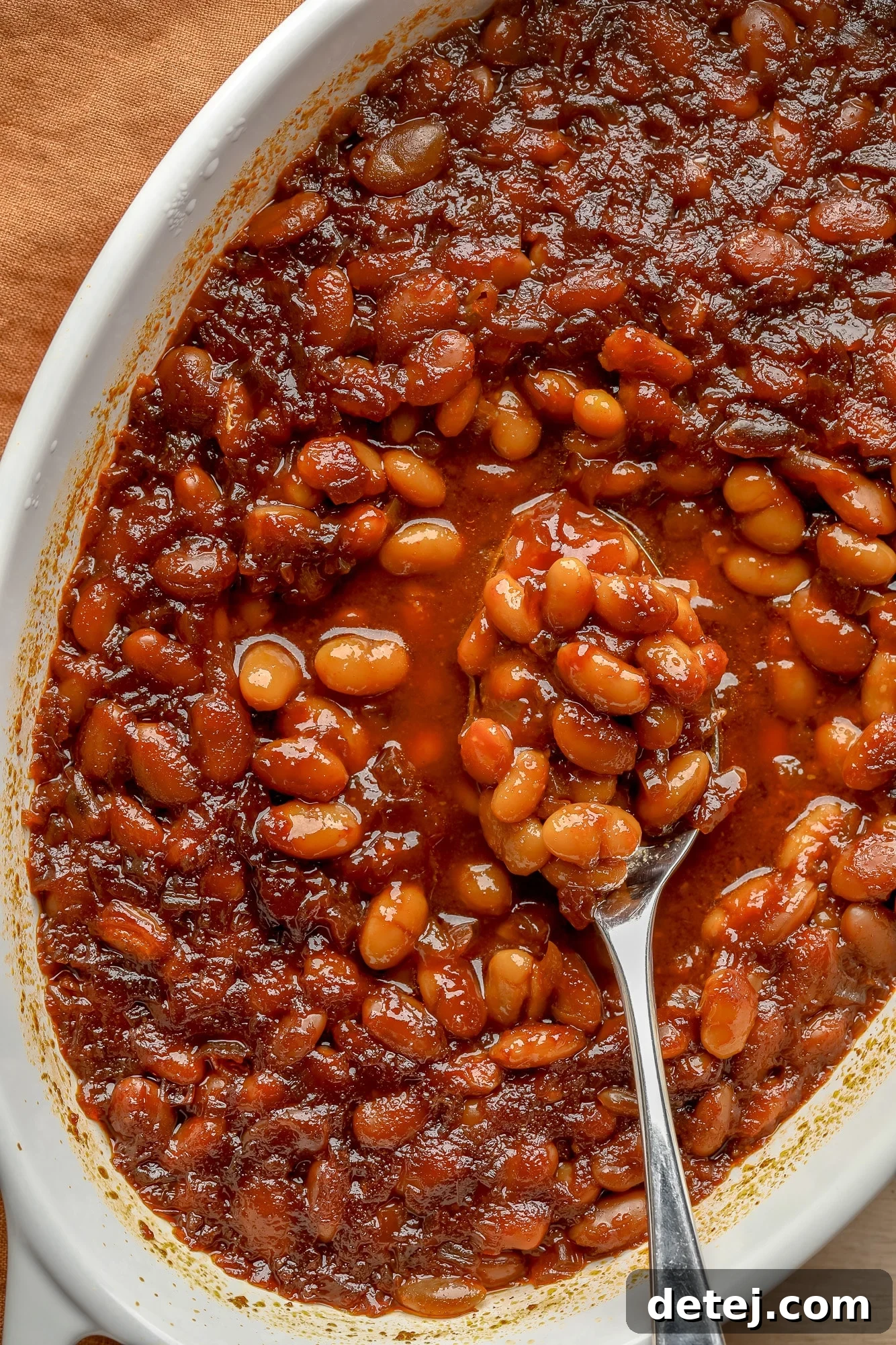 A spoon scooping a generous portion of hot, savory vegan baked beans from a casserole dish, highlighting their rich texture and thick sauce.