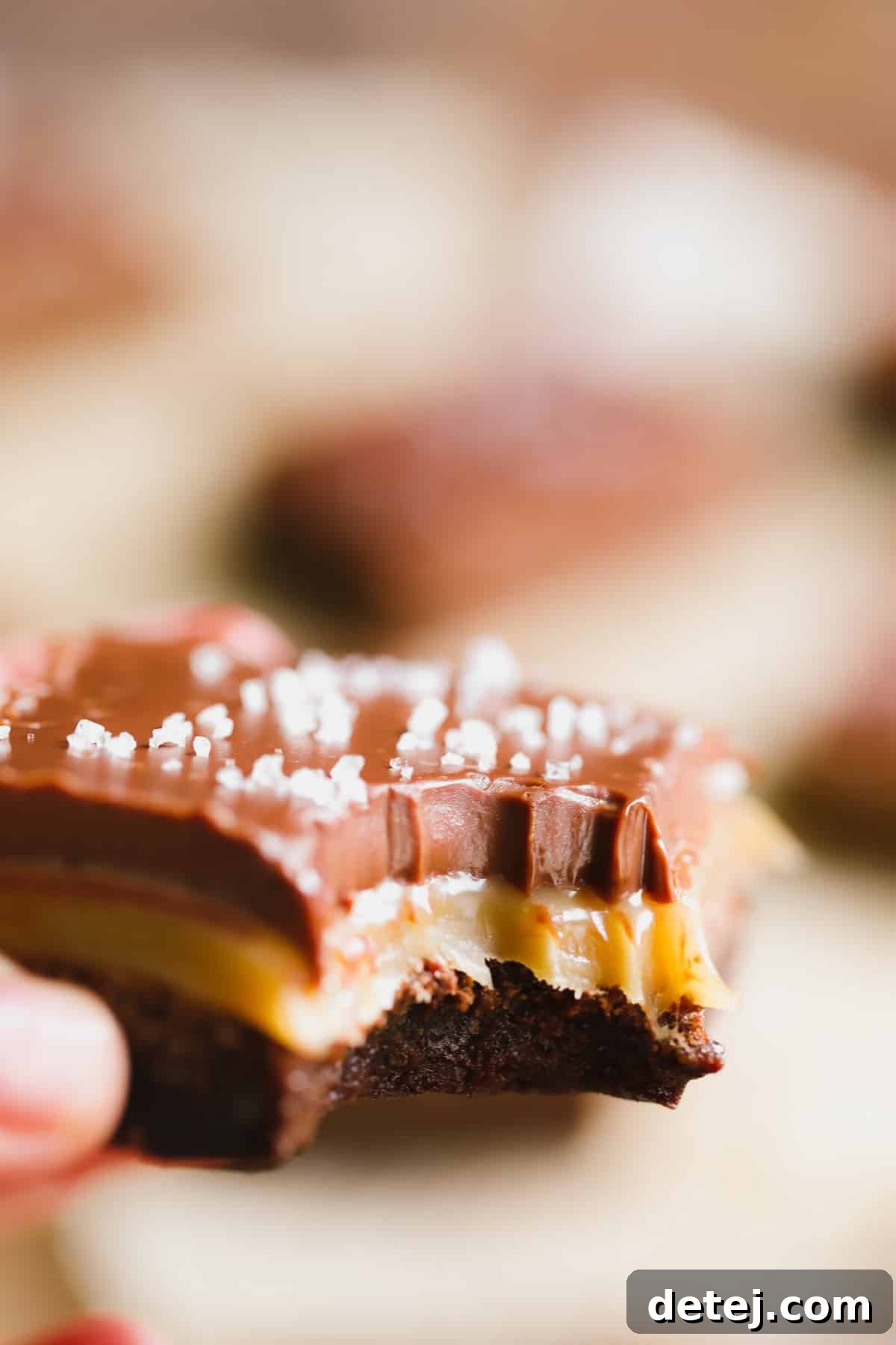 A perfectly baked vegan salted caramel brownie with a bite taken out, revealing the distinct layers of fudgy chocolate, gooey caramel, and smooth ganache. A hand holds the brownie, emphasizing its irresistible appeal.