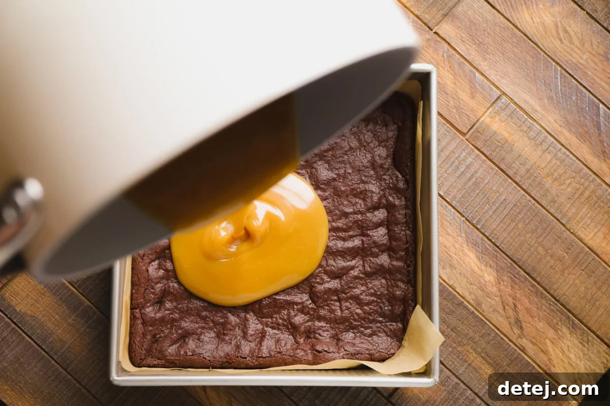 Thick, golden salted caramel being generously poured over the cooled chocolate brownies in a baking pan, creating an irresistible middle layer.