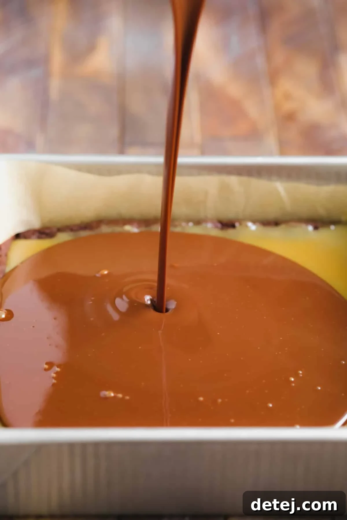 Smooth, dark chocolate ganache being poured over the set caramel layer on top of the brownies in a pan, completing the three-tier dessert.