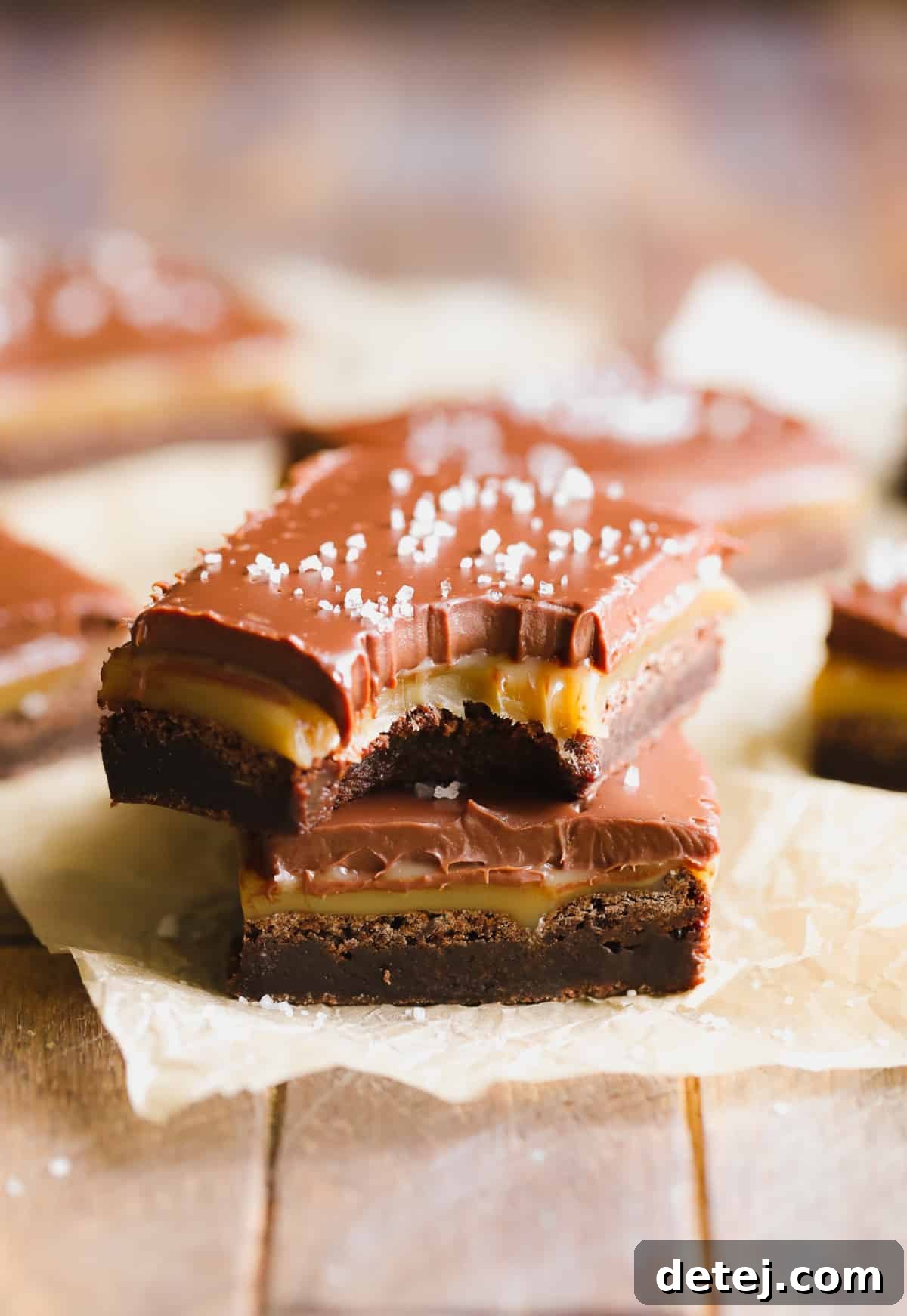 Two exquisitely crafted vegan salted caramel brownies, featuring thick layers of caramel and chocolate ganache, sprinkled with sea salt, resting on parchment paper. The details highlight their deliciousness.