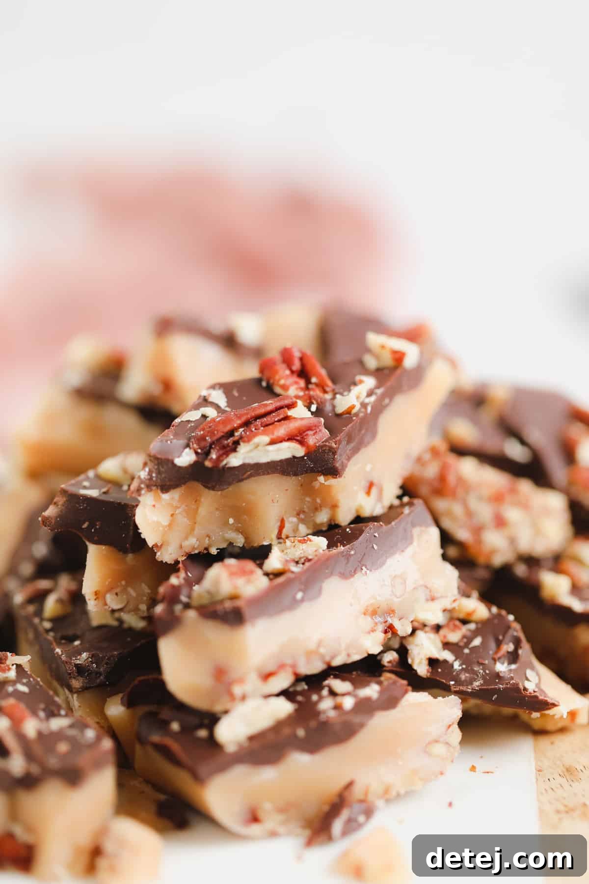 Vegan toffee squares topped with melted chocolate and chopped pecans, artfully stacked against a soft pink backdrop.