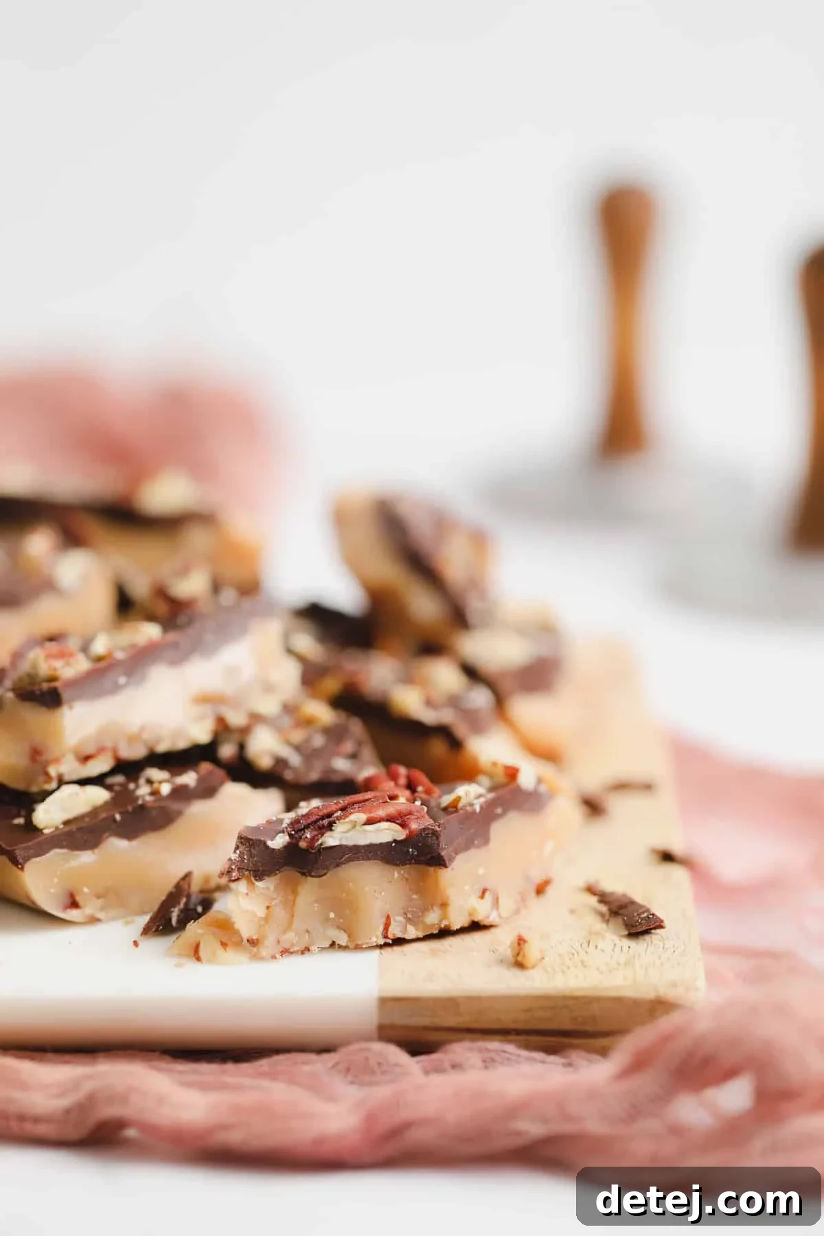 Close-up of golden vegan toffee pieces, some coated in chocolate and sprinkled with nuts, arranged on a white and wooden serving board.