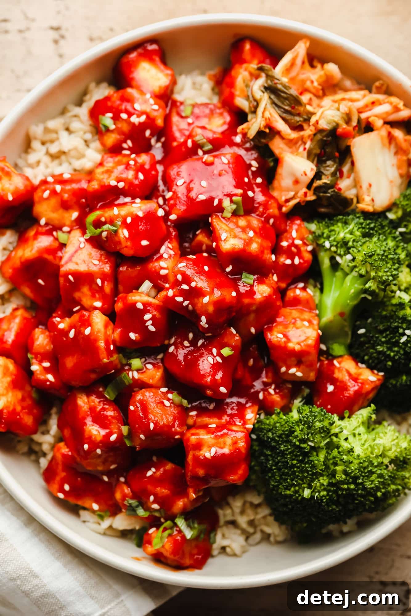 Close up on Gochujang Tofu in a white bowl with rice, broccoli, and kimchi, garnished with sesame seeds and green onions.