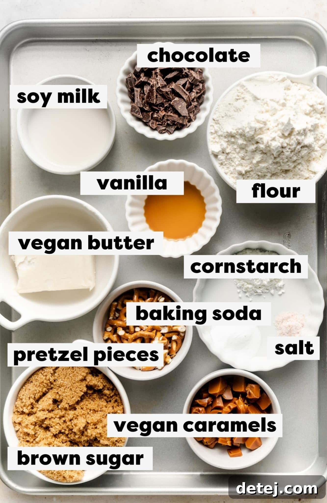An inviting overhead shot of various essential ingredients for vegan kitchen sink cookies neatly arranged on a pristine white surface, including softened vegan butter, packed brown sugar, all-purpose flour, rich chocolate, crunchy pretzels, and soft caramel pieces.