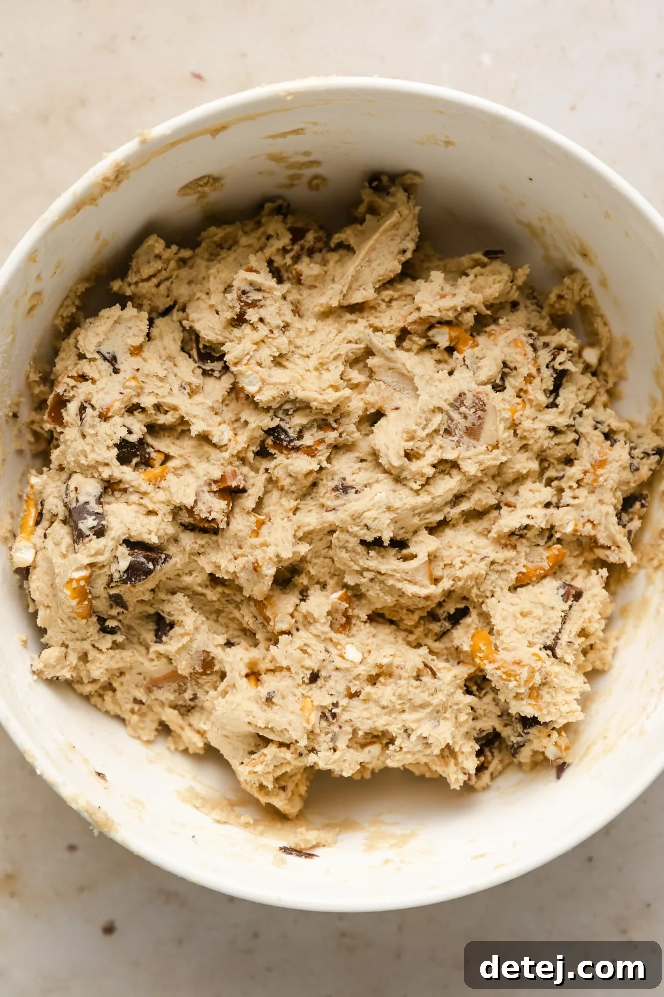 Raw vegan kitchen sink cookie dough in a white bowl, visibly studded with rich chocolate chunks, gooey caramel pieces, and crunchy crushed pretzels, ready for chilling.