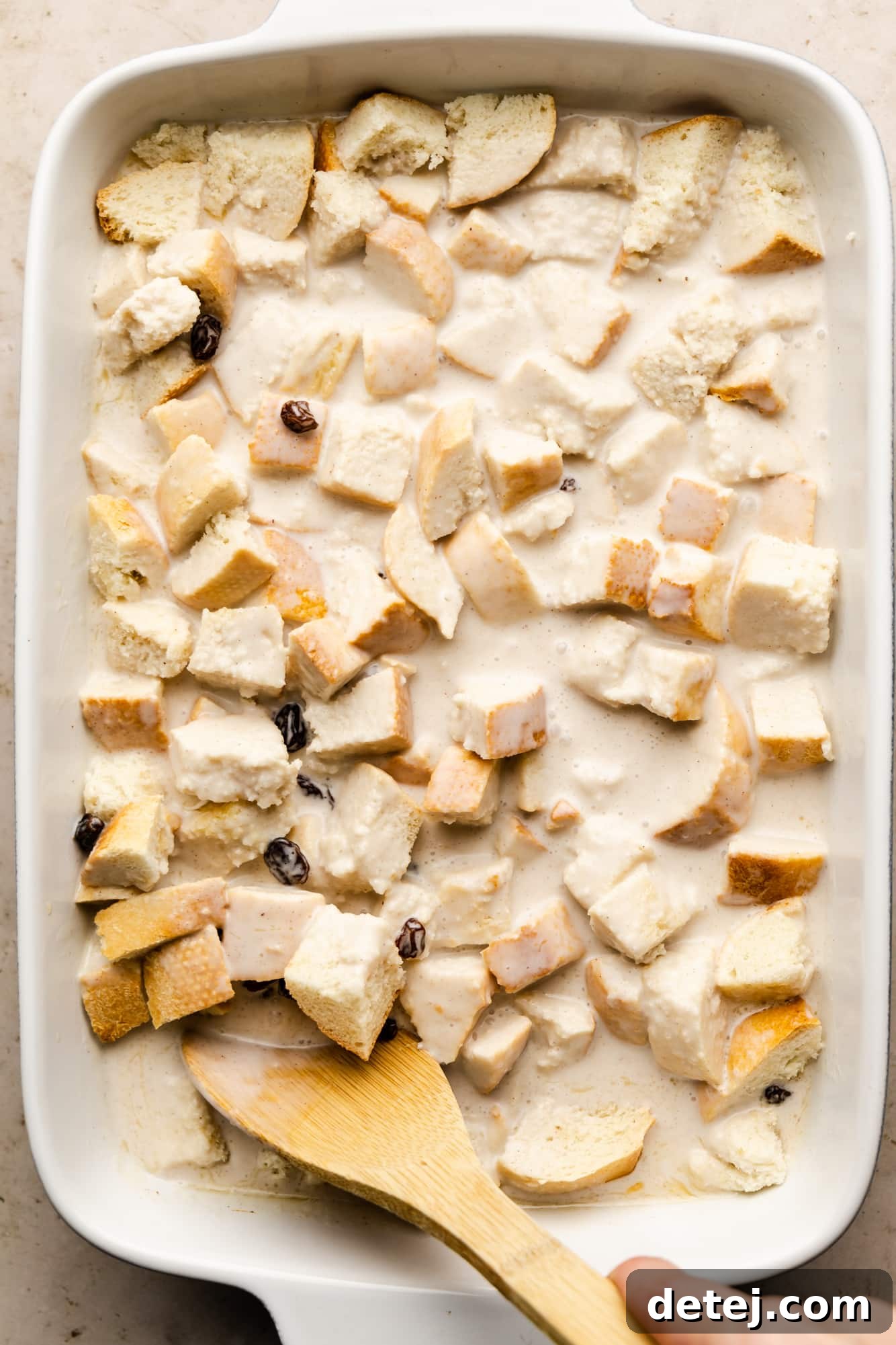 Decadent Vegan Bread Pudding 5 A woman's hand gently stirs cubed bread coated in a creamy white sauce with bourbon-soaked raisins in a white baking dish using a wooden spoon.