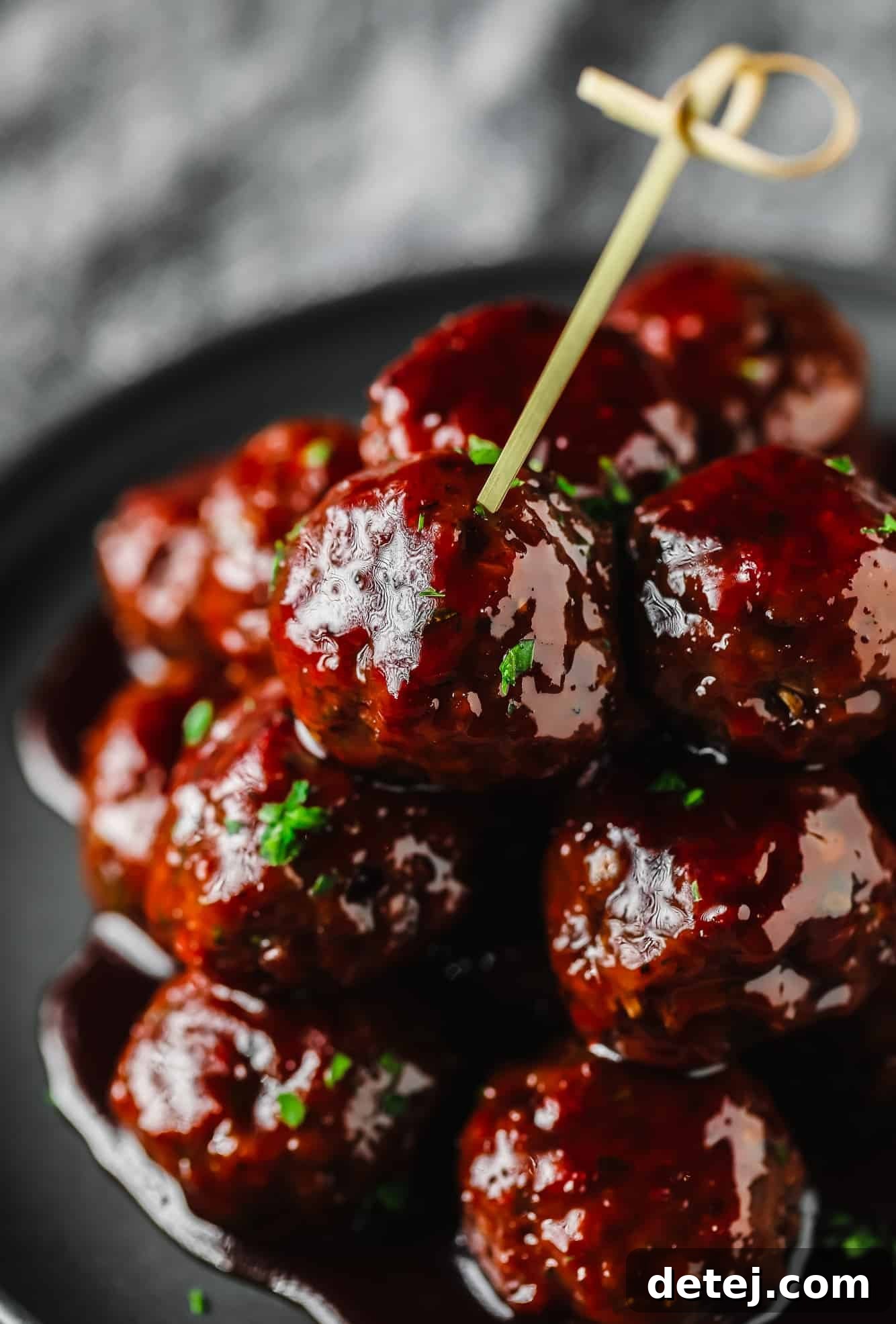 A vibrant pile of vegan grape jelly meatballs glistening with sauce on a grey plate, with a toothpick ready for serving.