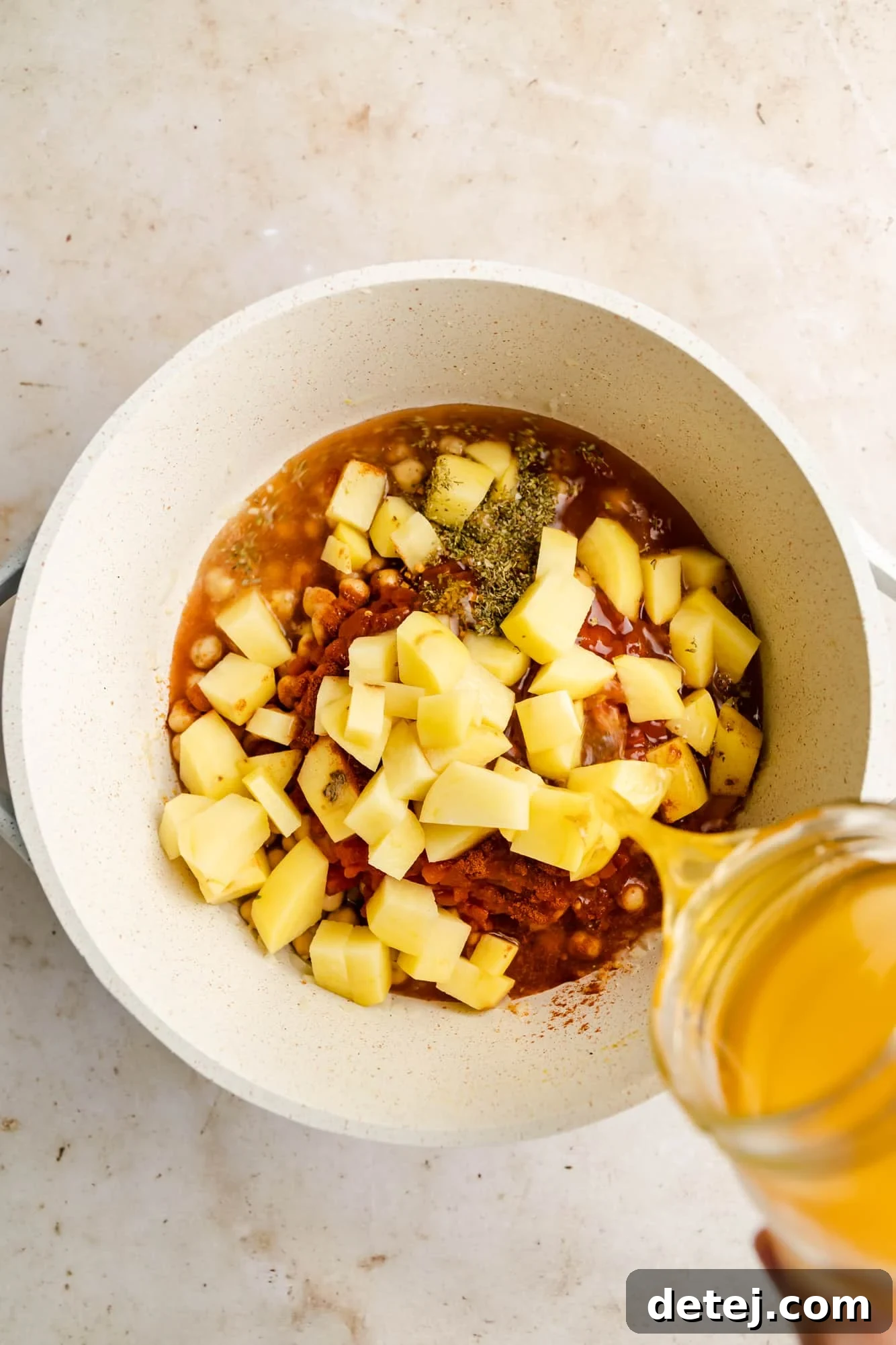 Pouring vegetable broth into a white pot filled with a vibrant mix of diced potatoes, chickpeas, tomatoes, and aromatic herbs.