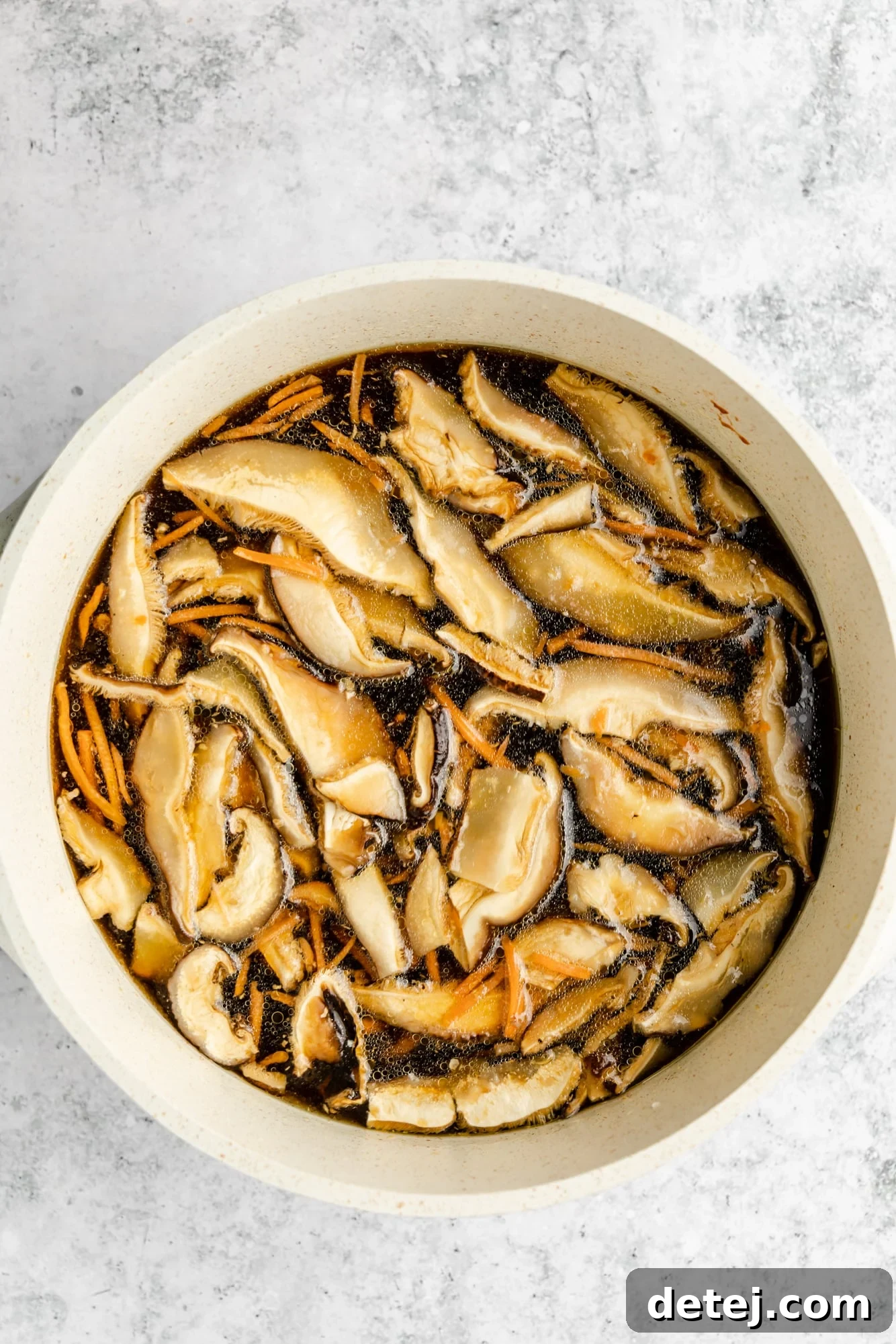 Mushrooms and shredded carrots visibly simmering in a rich, dark brown broth within a large white pot, indicating the flavors are melding.